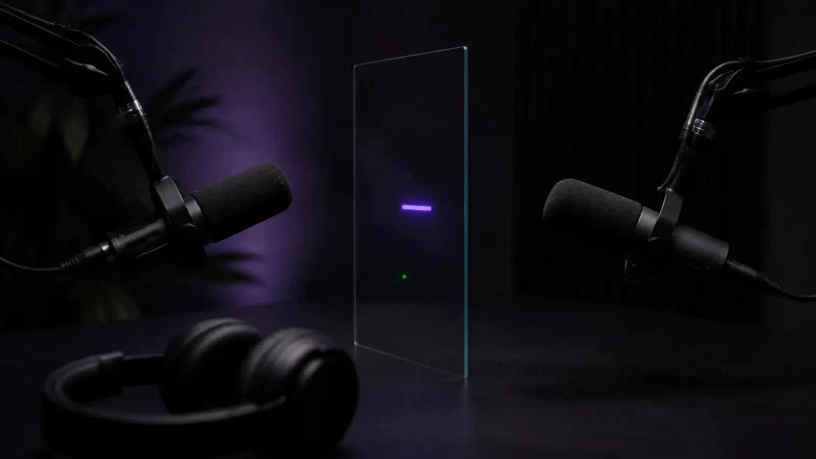 Two podcast microphones facing each other across a dark desk, a thin glass pane with a small glowing bar standing between them.