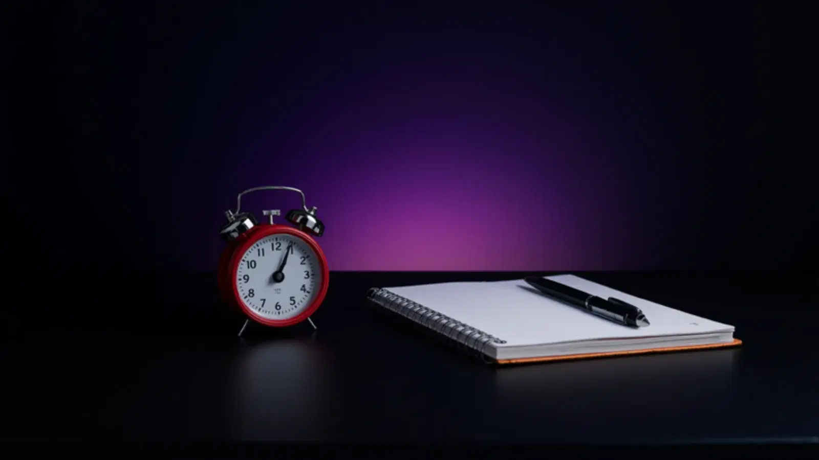 A small red alarm clock beside a notebook and pen on a dark desk.